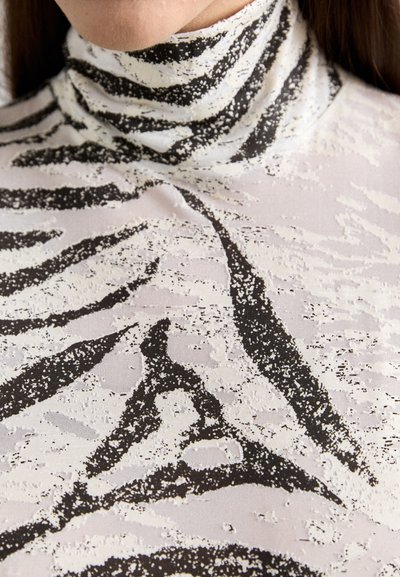 Majica s visokim ovratnikom, koja se ističe crno-bijelim uzorkom zebre, izrađena od glatkog, teksturiranog materijala. Istaknute tamne pruge na svijetloj pozadini.