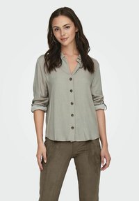 Camisa de botones de tela suave en un gris claro, con las mangas arremangadas, que presenta un cuello y cinco botones oscuros en la parte delantera.