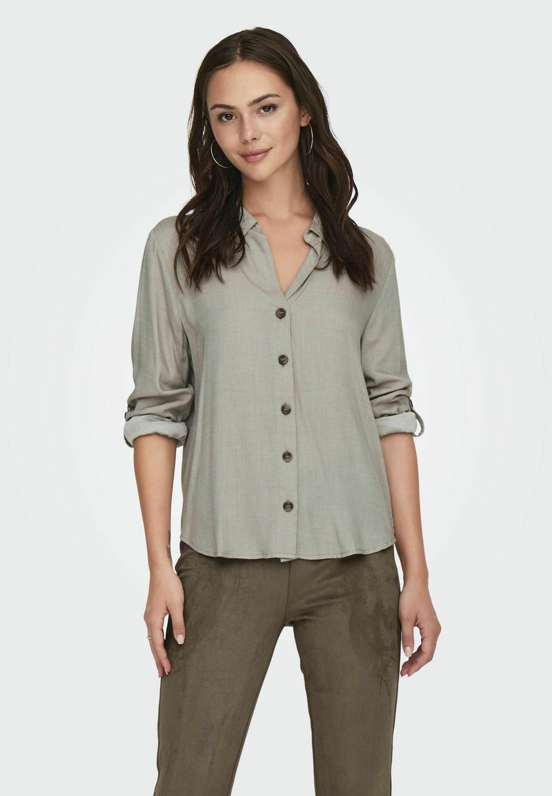 Camisa de botones de tela suave en un gris claro, con las mangas arremangadas, que presenta un cuello y cinco botones oscuros en la parte delantera.