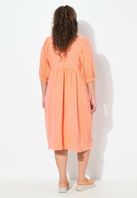 Vestido de color coral, a la altura de la rodilla, con mangas abullonadas, tejido texturizado y cintura fruncida. Llevado con sandalias de cuña en un tono gris claro.