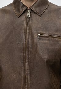 Gros plan d'une veste en cuir marron avec une fermeture éclair à l'avant et une poche poitrine zippée sur le côté droit.