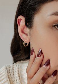 Pendiente de aro dorado retorcido usado en la oreja, con un acabado brillante. Uñas largas y puntiagudas de color burdeos tocan la piel cerca de la oreja.