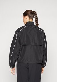 Zwarte windbreaker jack met een losse pasvorm, hoge kraag, elastische manchet en witte piping details langs de schouders en zijkanten.