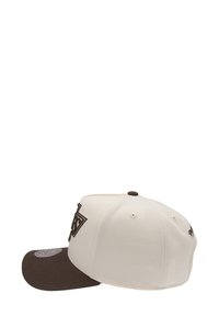 Casquette crème avec une visière marron, ornée d'un logo en relief marron foncé à l'avant. Conçue avec une construction à six panneaux et des oeillets.