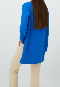 Blusa azul de manga larga con un corte holgado, textura sedosa y aberturas laterales, combinada con pantalones beige ajustados. Vista trasera, el cabello lacio cae hacia abajo.