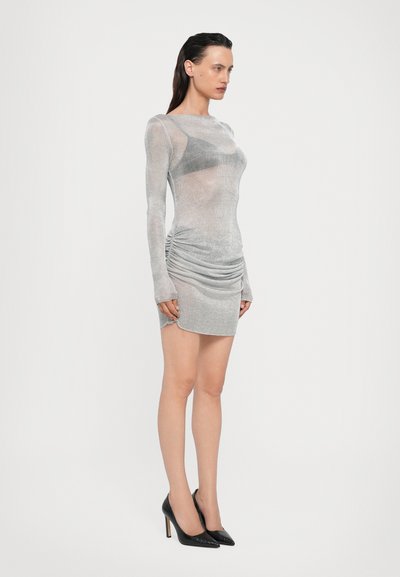 Vestido mini metálico plateado con mangas largas, detalles fruncidos en los laterales y un aspecto transparente, combinado con tacones negros de punta afilada.