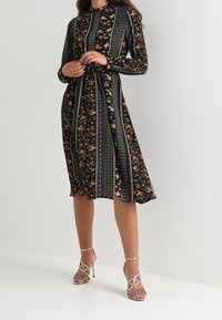 Robe noire à manches longues avec imprimé floral et détails rayés ; longueur genou avec taille cintrée et jupe fluide. Sandales à talons hauts argentées.