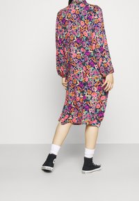 De bloemdeken heeft een donkere achtergrond met levendige roze, oranje en groene bloemen. Lange mouwen, losse pasvorm, gedragen met zwarte sneakers.