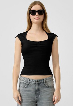 Kobieta w czarnej dopasowanej koszulce z krótkim rękawem i kwadratowym dekoltem, szare dżinsy z wysokim stanem oraz czarne okulary przeciwsłoneczne w kształcie kocich oczu, stojąca na tle białego tła.