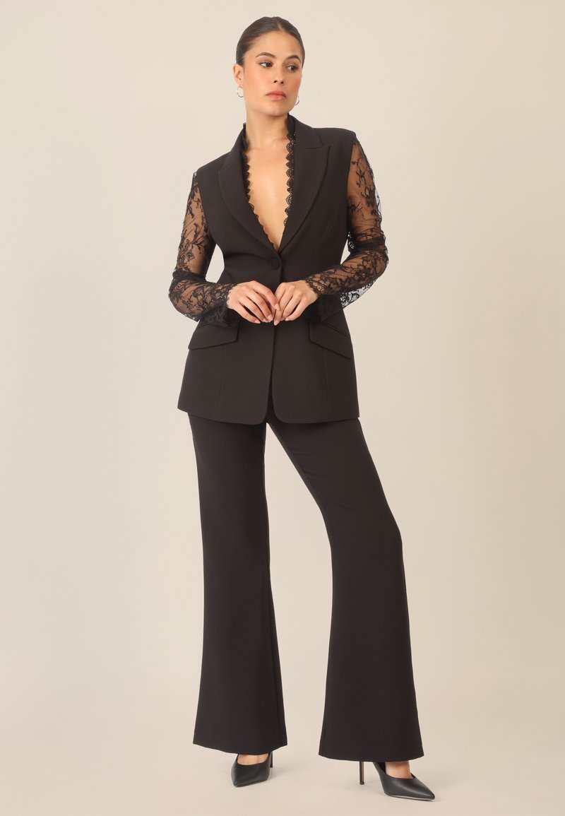 Schwarzer Hosenanzug mit einem maßgeschneiderten Blazer mit spitzendetaillierten Ärmeln, tiefem V-Ausschnitt und weit geschnittenen Hosen. Schwarze Absätze runden das Outfit ab.