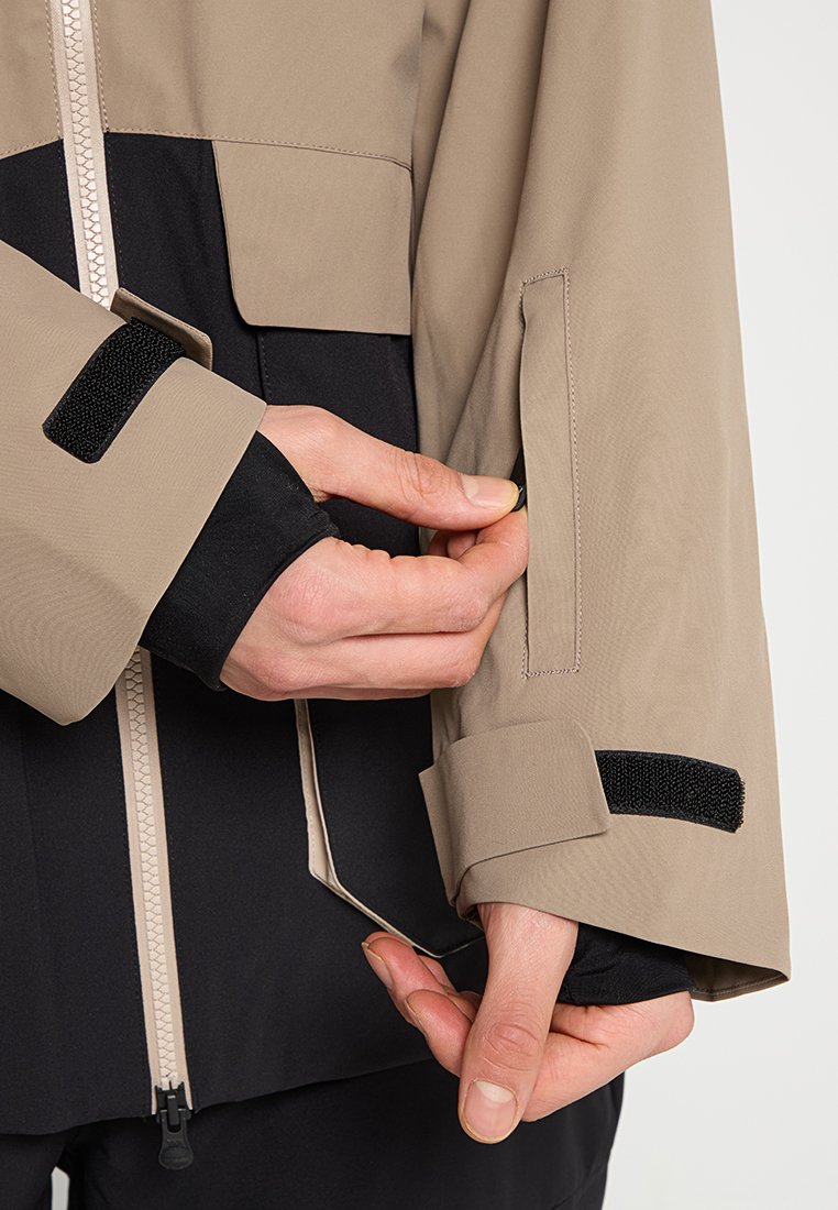 Hände, die den Beigefarbenen Jackenärmel mit schwarzen Klettverschlüssen und einem Reißverschluss über einem schwarzen Langarmoberteil befestigen.