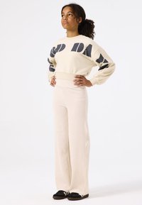 Creme sweatshirt met grote zwarte letters, gecombineerd met lichtbeige brede ribbroek. Zwarte sneakers maken de outfit af.