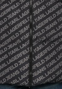 Juodas pūkinis švarkas su pasikartojančiu pilku "KARL LAGERFELD JEANS" logotipo spaudiniu, turintis centrinius užtrauktuką ir lygų, blizgų audinio tekstūrą.