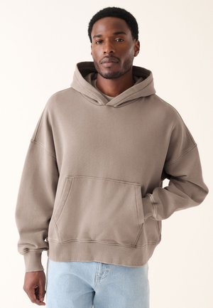 Uomo con capelli corti e ricci che indossa una felpa oversize color taupe e jeans azzurri chiari, in piedi davanti a uno sfondo chiaro e semplice.