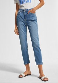 Vaqueros de tiro alto en un azul claro con un corte ajustado, que cuentan con un diseño clásico de cinco bolsillos y un dobladillo cortado. Combinados con una camiseta blanca casual y sandalias.