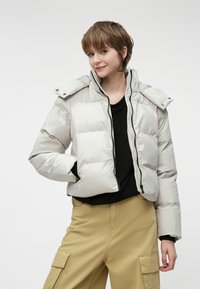 Jasnoszara kurtka puchowa z zapięciem na zamek, kołnierzem na napy i elastycznymi mankietami. Ma krótki krój i subtelny połysk; zestawiona z beżowymi spodniami.