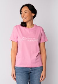 Rosa Baumwoll-T-Shirt mit kurzen Ärmeln und rundem Halsausschnitt, mit weißem Text auf der Vorderseite. Blaue Jeans vervollständigen das Outfit.
