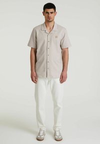 Camisa de manga corta y cuello en beige claro con rayas finas, que presenta un bolsillo en el pecho izquierdo y pantalones blancos de corte relajado con zapatillas.