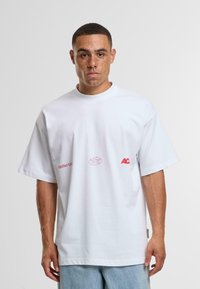 Bílý bavlněný tričko s krátkým rukávem, s červenými logy na hrudi. Kolo výstřih, uvolněný střih a měkká textura.