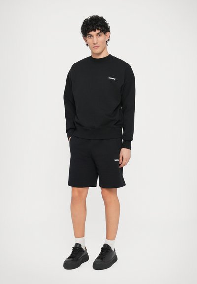 Ensemble sweat-shirt noir et short, avec un col rond et un petit logo. Le tissu semble doux, avec une texture lisse. Des baskets noires complètent le look.