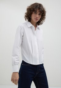 Camicia bianca a maniche lunghe con colletto classico, caratterizzata da una vestibilità su misura e una texture liscia, infilata in jeans blu scuro.