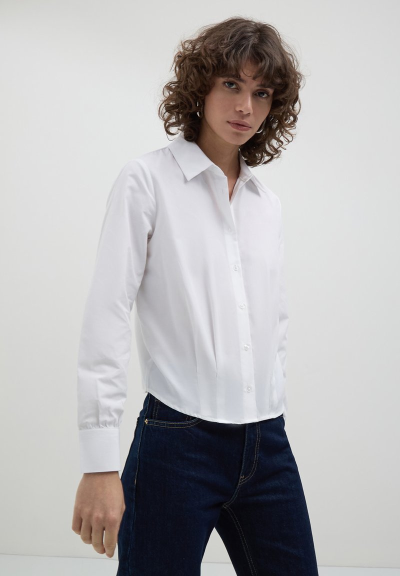 Camicia bianca a maniche lunghe con colletto classico, caratterizzata da una vestibilità su misura e una texture liscia, infilata in jeans blu scuro.