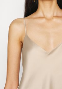 Camiseta de satén beige con finas tirantes tipo spaghetti, con escote en V y una textura suave y lisa, ideal para combinar o para un look casual.