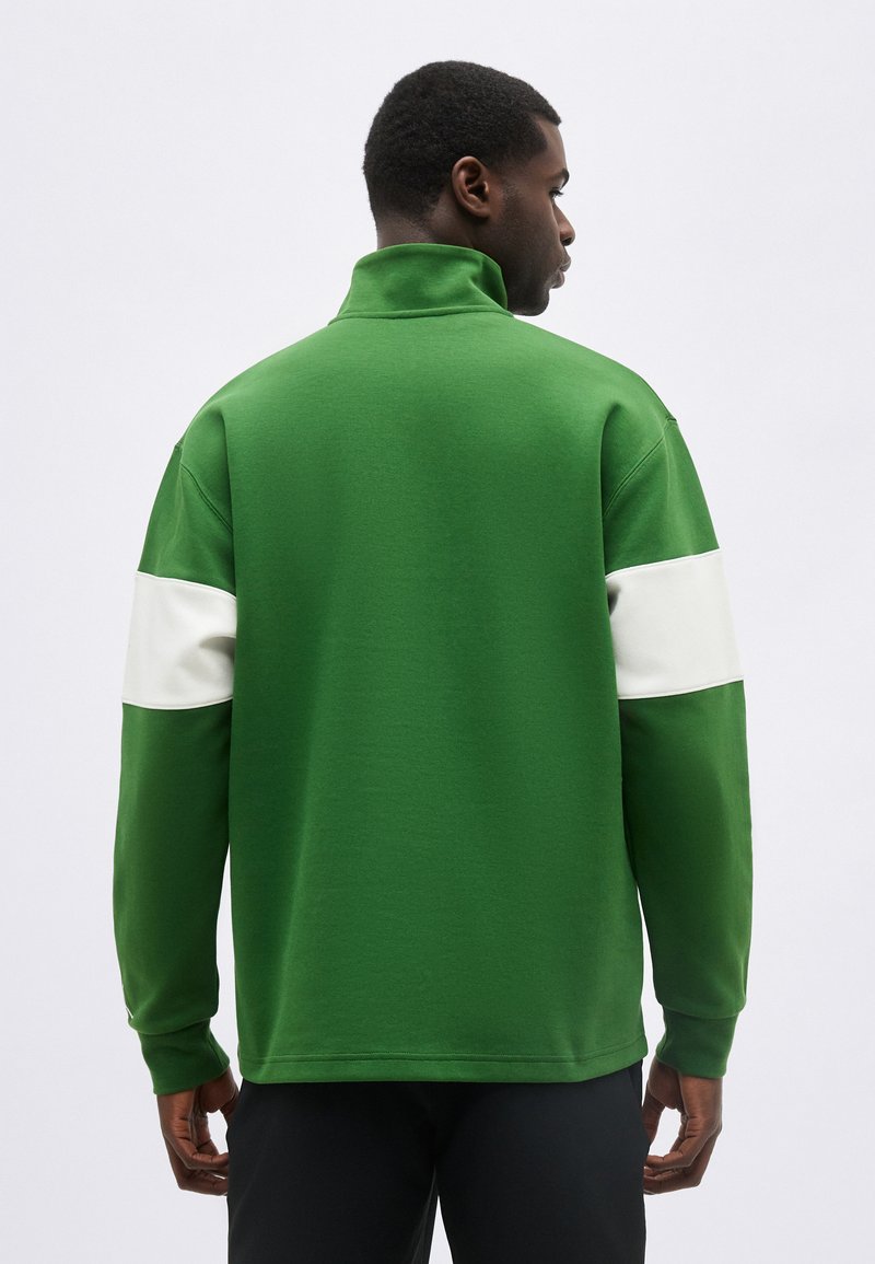 Homme vu de dos portant un haut vert à manches longues avec des rayures horizontales blanches sur le haut des bras et un pantalon noir.