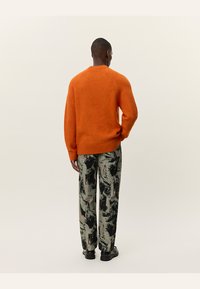 Uomo di spalle che indossa un maglione arancione, pantaloni astratti con motivo nero e beige, e scarpe nere contro uno sfondo chiaro uniforme.