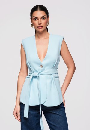 Kobieta w bezrękawowej, jasnoniebieskiej marynarce z paskiem i głębokim dekoltem w kształcie litery V oraz w ciemnoniebieskich dżinsach, stojąca na jednolitym tle.