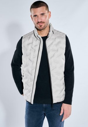 Man with light gray quilted vest over black long-sleeve shirt and blue jeans standing against light background.