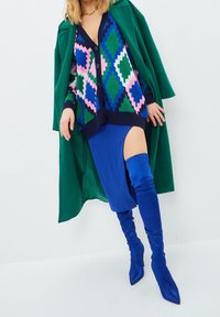 Colorful knit cardigan with geometric patterns, navy trim, and buttons, paired with a blue high-slit skirt and shiny blue thigh-high boots.