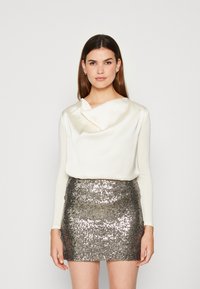 Weiße Bluse mit langen Ärmeln und drapiertem Ausschnitt, kombiniert mit einem metallisch silbernen Pailletten-Minirock mit glatter Textur und schimmernder Oberfläche.