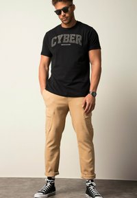 Schwarzes Baumwoll-T-Shirt mit "CYBER UNDERGROUND" Grafik, kombiniert mit beigen Cargo-Hosen und schwarzen High-Top-Sneakern mit weißen Akzenten.