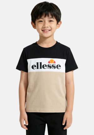 Jeune garçon souriant, portant un t-shirt ellesse noir, blanc et beige, debout devant un fond uni gris clair.