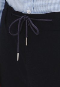 Ciemnoniebieskie spodnie z teksturowanej powierzchni, z zawiązywanym sznurkiem wykończonym srebrnymi detalami oraz w kratkę koszulą w tle.