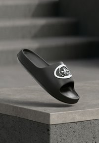 Juoda Adidas šlepetė su balto logotipo detalėmis, padėta ant betono krašto priešais neryškias laiptų pakopas.