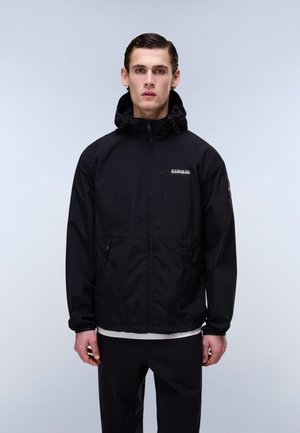 Ung mand iført en sort hættejakke med lynlås og sorte bukser, stående foran en ensfarvet lysegrå baggrund.