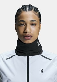 Black neck warmer made of soft fabric, featuring a slim fit. Worn with a white jacket, which has black accents and a zipper.