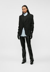 Čierny na mieru šitý blejzer cez čiernu vestu, svetlomodrá košeľa, kožené rukavice a priliehavé čierne nohavice. Lesklá textura, ostrý dizajn a štíhla silueta.