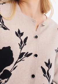 Beige stickad cardigan med svart blommönster, rund halsringning och fem svarta knappar med silverdetaljer. Mjuk textur.
