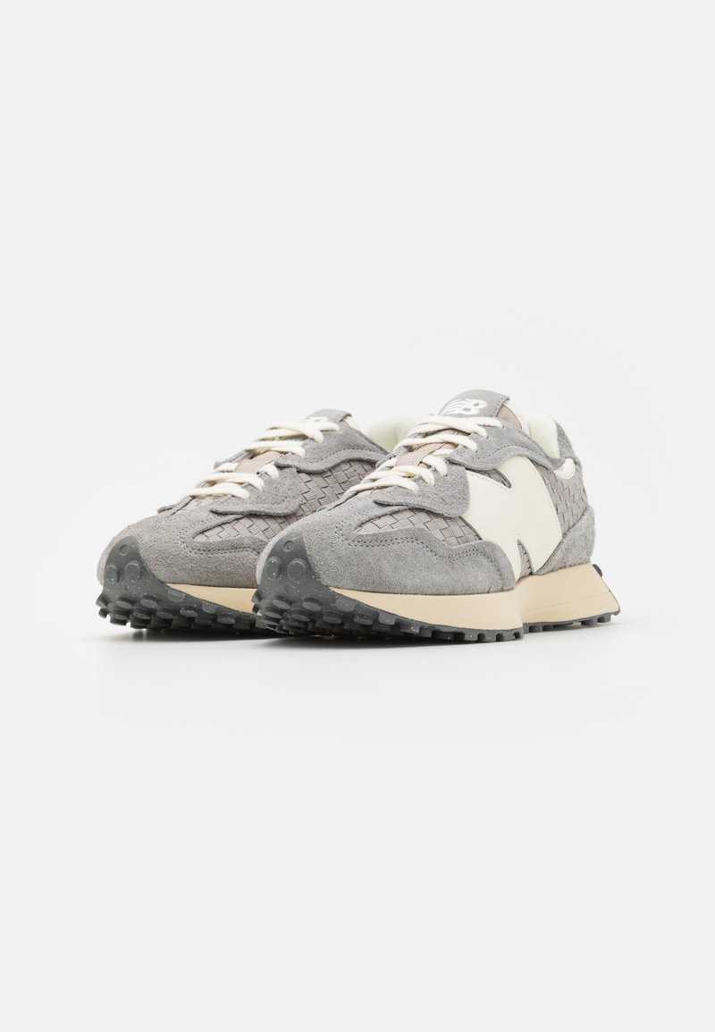 Un par de zapatillas texturizadas en gris y beige con cordones blancos y suelas gruesas de goma negra sobre un fondo blanco.