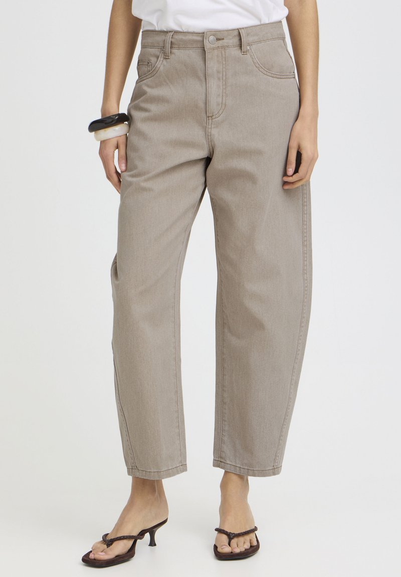 Personne portant un jean beige à jambes larges, un haut blanc, des bracelets noirs et blancs, et des sandales à talons noires avec des lanières tressées.