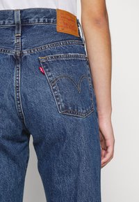 Niebieskie dżinsy Levi's z brązową skórzaną łatą, zakrzywionymi tylnymi kieszeniami z detalami w postaci szwów oraz czerwoną metką koło prawej kieszeni.