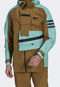 Veste imperméable bicolore marron et bleu clair avec multiples poches, fermeture éclair noire, sangle noire et rayures noires sur la manche.