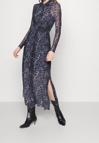 Langes, durchsichtiges Kleid mit Leopardenmuster in Lila- und Schwarztönen. Verfügt über lange Ärmel, einen durchgehenden Kragen und seitliche Schlitze. Wird mit schwarzen Stiefeln getragen.