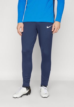 Mężczyzna w granatowych legginsach Nike, niebieskiej bluzie z długim rękawem i białych korkach stoi na jednolitym tle.