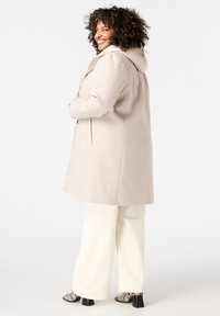 Beige wollen jas met een capuchon en subtiele textuur, met een getailleerde pasvorm, knoopsluiting en zakken aan de zijkant, gecombineerd met witte broek.