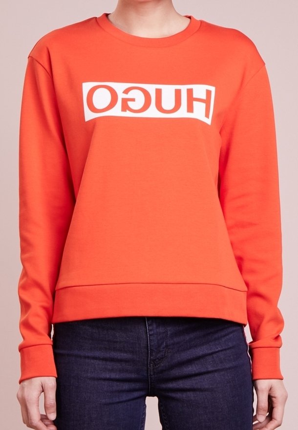 Personne portant un sweat-shirt à manches longues orange vif avec un texte blanc en miroir "HUGO" sur la poitrine et un jean bleu foncé sur un fond uni.