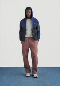 Marineblaue und schwarze Nike-Kapuzenjacke mit Reißverschluss und lila Logo, graues Shirt darunter und locker sitzende dunkelrote Hose. Modell trägt Turnschuhe auf blauem Boden.